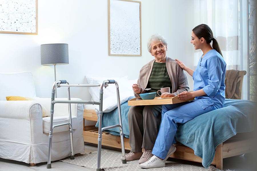 商业保险-高级护理设施，一个快乐的女人坐在她的床上，护士把手放在她的肩膀上