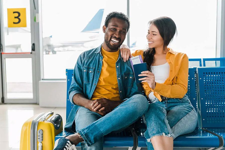 Travel Insurance - Smiling Young Couple Sitting in the Departure Lounge with Baggage and Tickets in the Airport with Airplane behind in the Distance